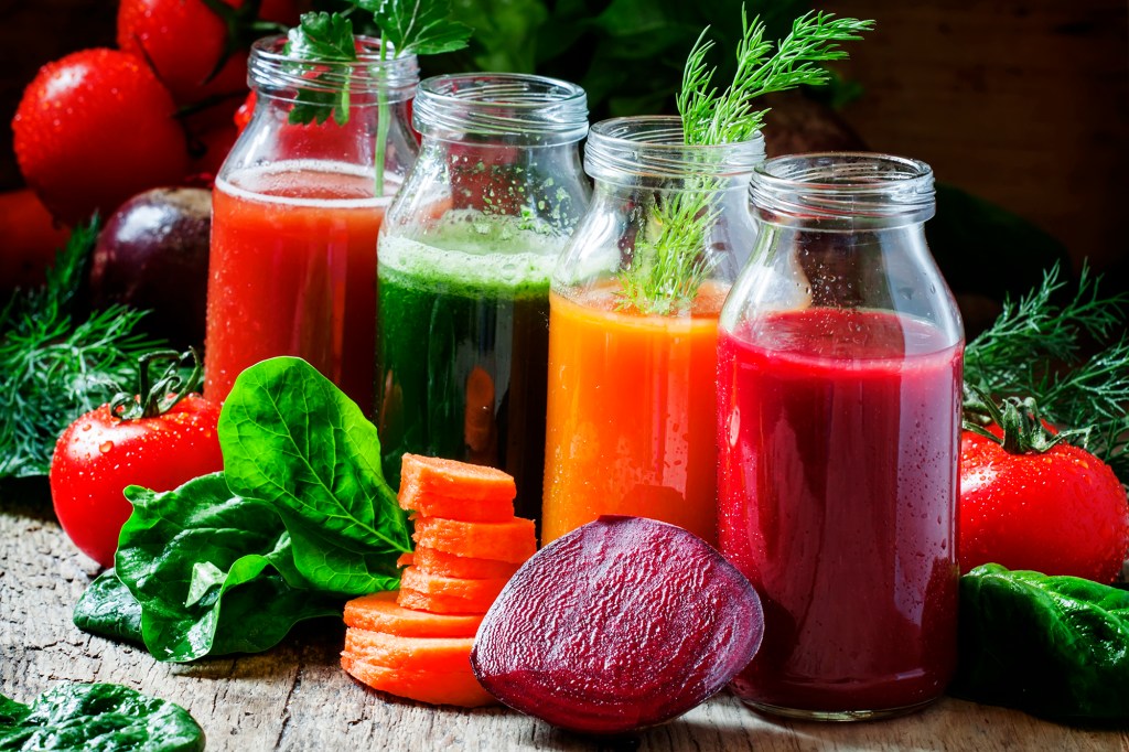 Four kind of vegetable juices: red, burgundy, orange, green, in small glass bottles, fresh vegetables and herbs, vintage wooden background, selective focus